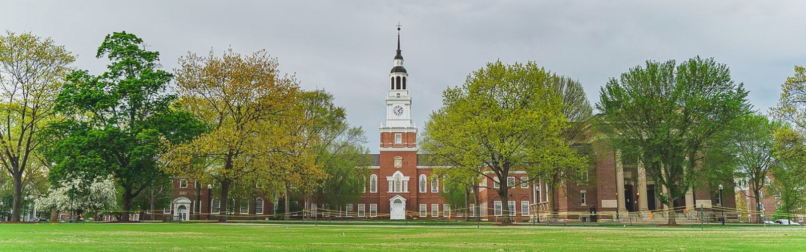 Image shows a brick building with a tall clock tower surrounded by trees on a green lawn under a cloudy sky.