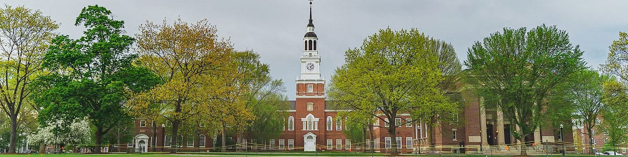Image shows a brick building with a tall clock tower surrounded by trees on a green lawn under a cloudy sky.