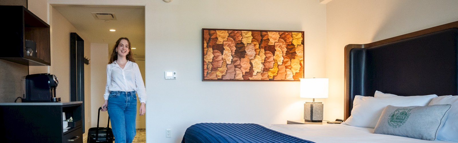 A woman is entering a modern hotel room with a neatly made bed, a blue blanket, wall art, and a small kitchenette on the left.