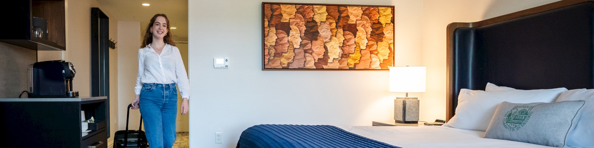 A woman is entering a modern hotel room with a neatly made bed, a blue blanket, wall art, and a small kitchenette on the left.