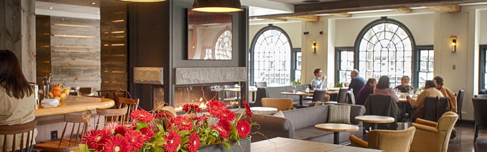 A cozy cafe with wooden tables, decorative red flowers, hanging lights, and people seated at tables near large arched windows.