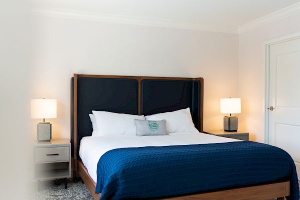 The image shows a neatly made bed with dark blue bedding, flanked by two nightstands, each with a lamp. The room has a clean, modern decor.