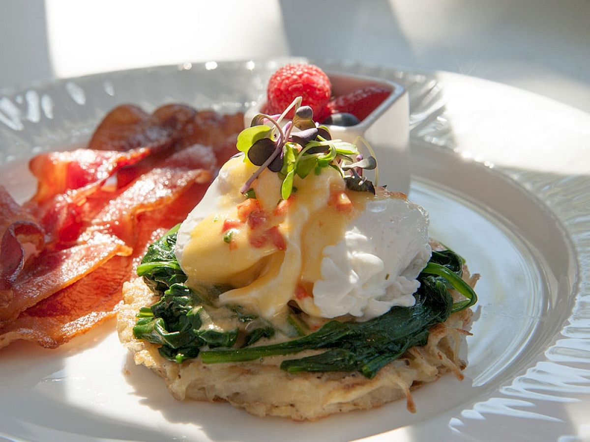 A beautifully plated brunch dish featuring poached eggs on spinach with hollandaise sauce, crispy bacon, and fresh strawberries.
