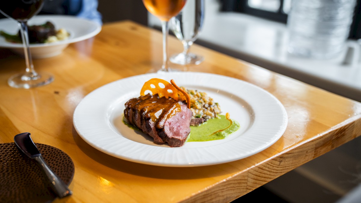 A gourmet plate with sliced meat, green sauce, and garnish on a wooden table, accompanied by two glasses of wine.