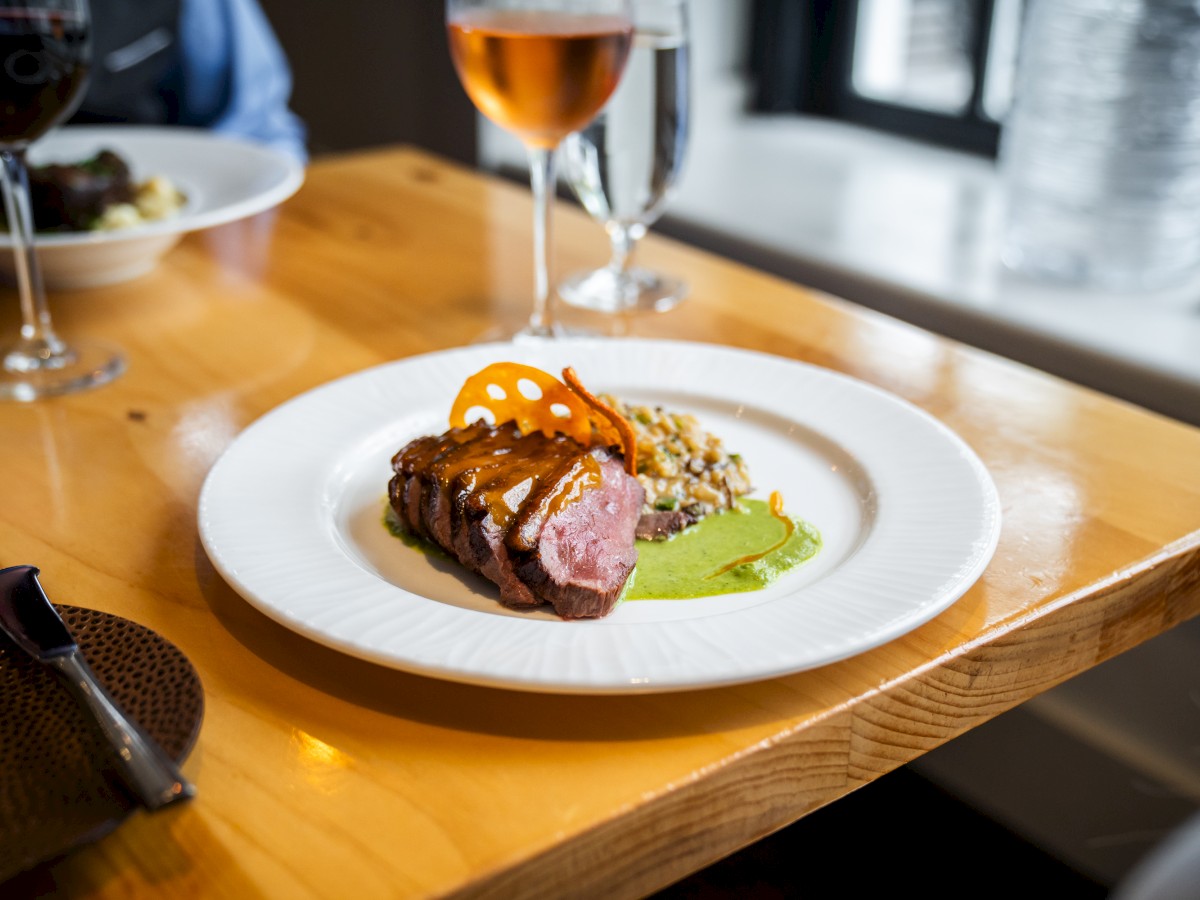 A gourmet plate with sliced meat, green sauce, and garnish on a wooden table, accompanied by two glasses of wine.