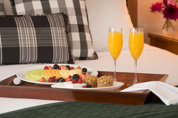 A breakfast tray on a bed with sliced fruits, berries, muffins, and two glasses of orange juice. Pillows, a green blanket, and flowers are visible.