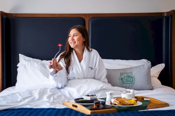 A person in a white robe is sitting on a bed with a breakfast tray, smiling while holding a fork with fruit.