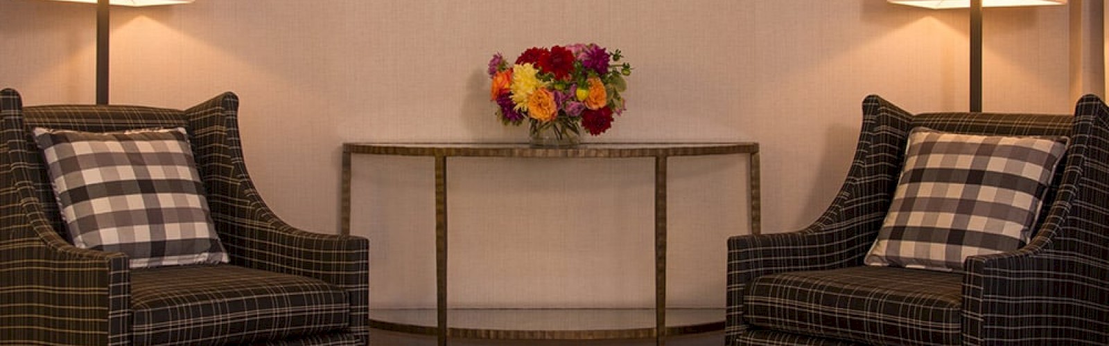 The image shows a cozy seating area with two armchairs, a console table with a vase of flowers, two table lamps, and framed pictures on the wall.
