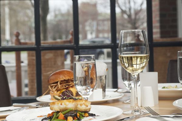 A table with a burger, vegetables, water, and wine set near large windows in a restaurant, with an outdoor scene visible.