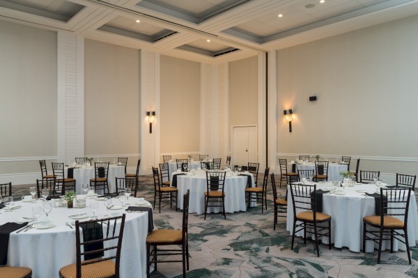 The image shows an elegant banquet room with round tables set with white tablecloths and black chairs, ready for an event or gathering.