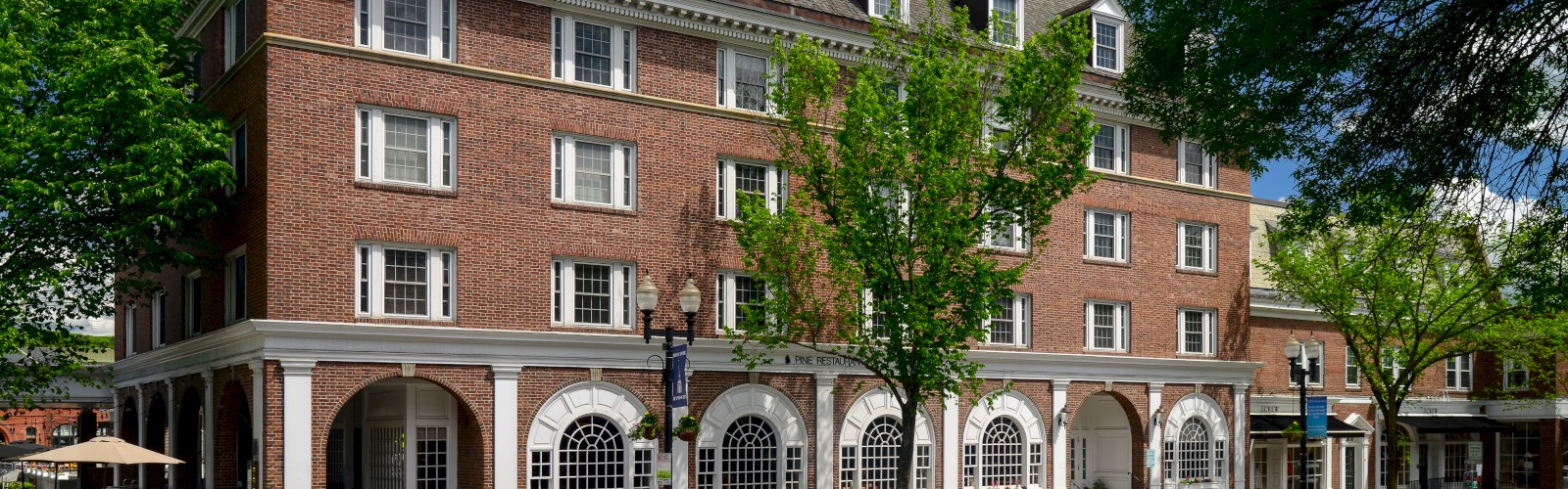 A four-story red brick building with arch windows and outdoor seating on the sidewalk, surrounded by trees and a clear blue sky.