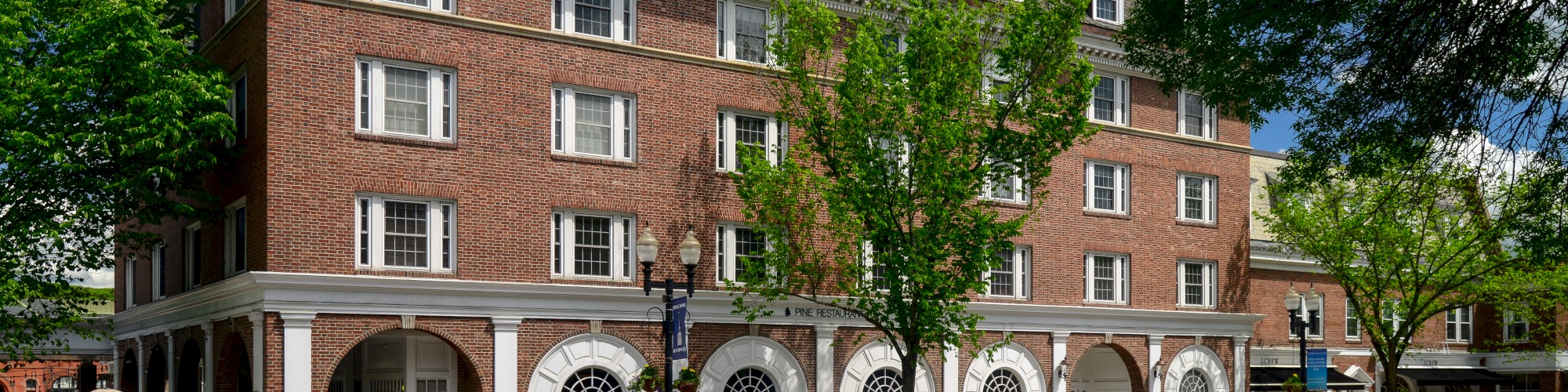 A four-story red brick building with arch windows and outdoor seating on the sidewalk, surrounded by trees and a clear blue sky.