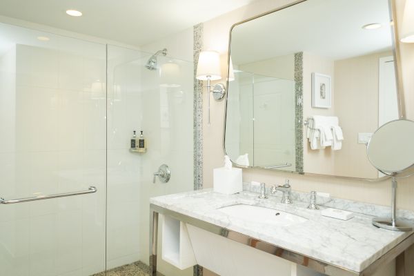 The image shows a modern bathroom with a glass shower, marble countertop sink, large mirror, wall-mounted toiletries, and neatly arranged towels.