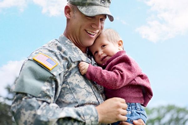 A soldier in uniform smiles while holding a small child who is resting their head on the soldier's shoulder outside under a blue sky.