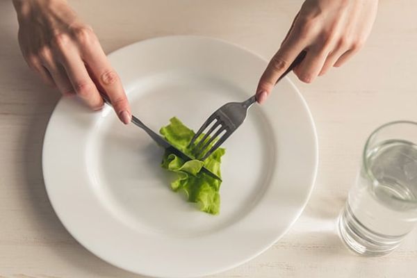 A person using a fork and knife to cut a small portion of lettuce on a white plate, next to a glass of water.