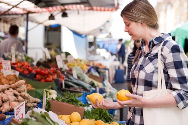 A woman is shopping at an outdoor market, holding two lemons and looking at produce, with various vegetables and fruits displayed on the stalls around her.