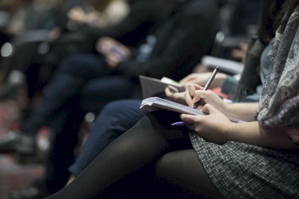 People are seated and taking notes with pens and notebooks, suggesting a conference, seminar, or workshop environment.