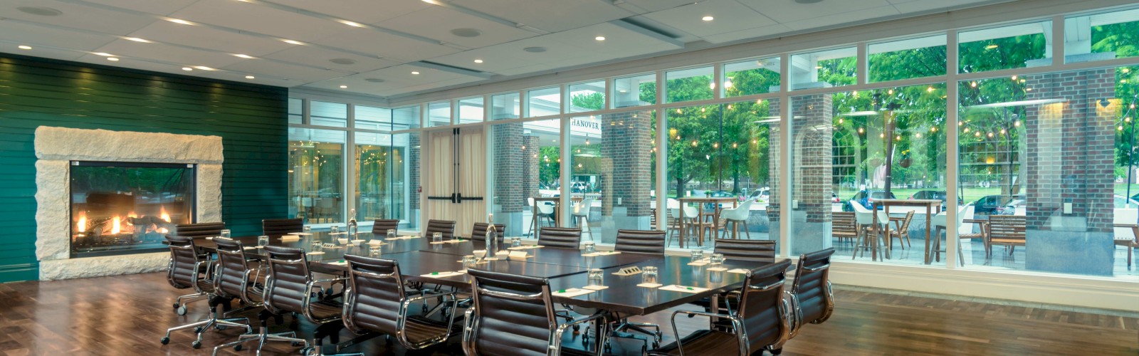 A modern conference room with a large table, black chairs, a fireplace, and large windows showing an outdoor patio with string lights.