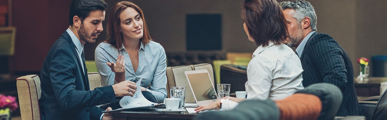 Four people in business attire are having a meeting in a casual setting, with papers, a laptop, and coffee cups on the table.