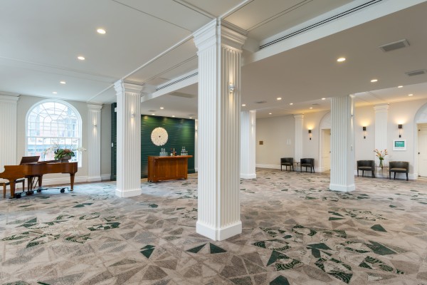 A spacious lobby with pillars, patterned carpet, a piano, chairs, a decorative wall, and a beverage station, well-lit and elegantly designed.