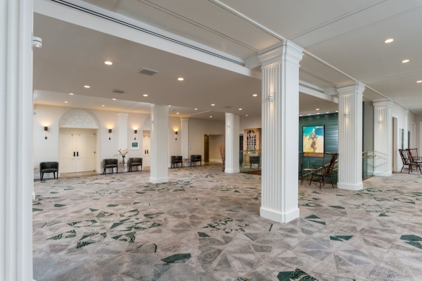 A spacious lobby with white columns, patterned carpet, chairs, and wall art, featuring a modern and elegant design.