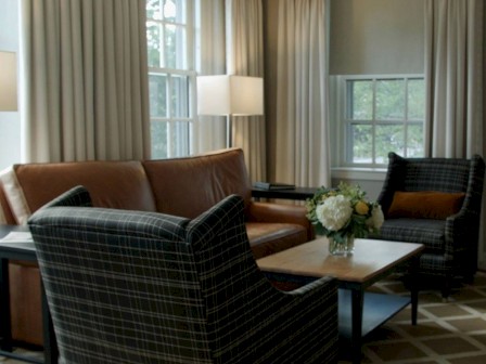 A cozy living room with a leather sofa, two checkered armchairs, a wooden coffee table with a vase of flowers, and tall windows with curtains.