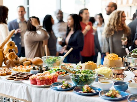 A diverse selection of food is displayed on a table in the foreground, while a group of people interacts in the background at a social gathering.