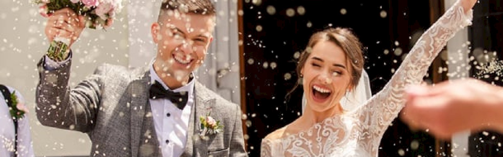A bride and groom are celebrating their wedding, surrounded by confetti, with the groom holding a bouquet and the bride raising her arm joyfully.