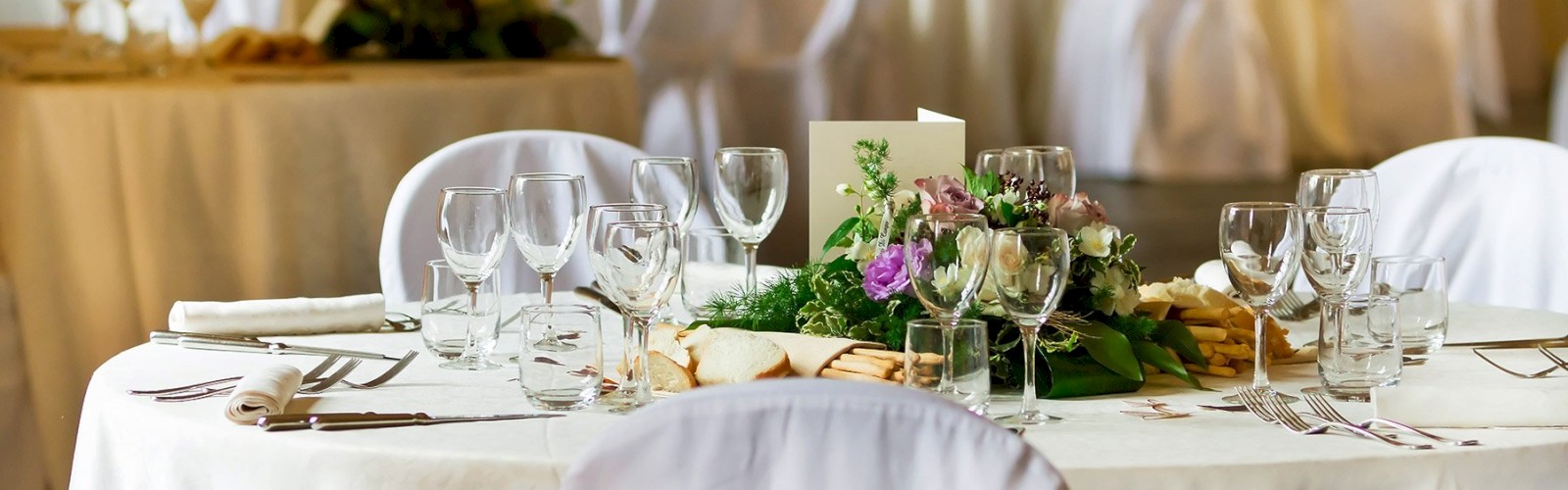 A round table is elegantly set with multiple glasses, silverware, a floral centerpiece, and napkins. The scene suggests a banquet or wedding event.