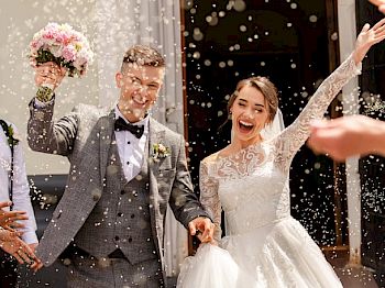 A newlywed couple is joyfully celebrating outside, with the groom holding a bouquet and both smiling amidst a shower of confetti.