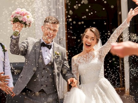 A newlywed couple is joyfully celebrating outside, with the groom holding a bouquet and both smiling amidst a shower of confetti.