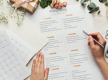 A person is reviewing a wedding planning checklist with a calendar, flowers, rings, pen, and smartphone on a table, while checking off tasks with a pen.