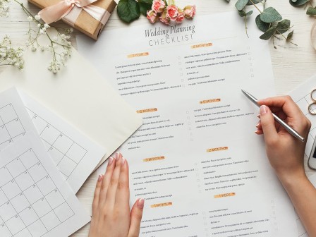 A person is reviewing a wedding planning checklist with a calendar, flowers, rings, pen, and smartphone on a table, while checking off tasks with a pen.