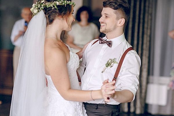 A bride and groom are dancing, smiling at each other tenderly. The bride wears a white dress and veil while the groom wears a shirt and bow tie.