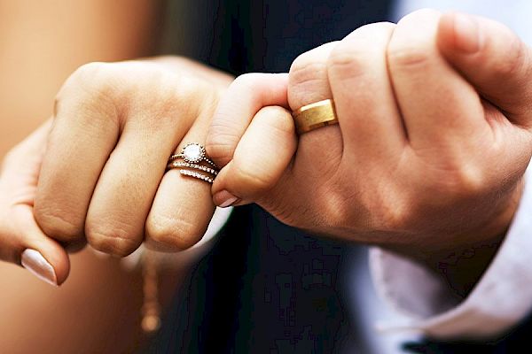 A close-up image of two hands with wedding rings, pinky fingers linked together, showing signs of a commitment or marriage ceremony.