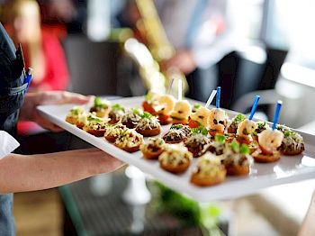 A person is holding a tray of bite-sized appetizers garnished with herbs, with blurred background elements including people and possibly a saxophone player.