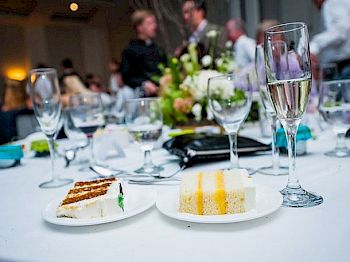 A table with slices of cake, an empty plate, wine glasses, and a bouquet, with people conversing in the background.