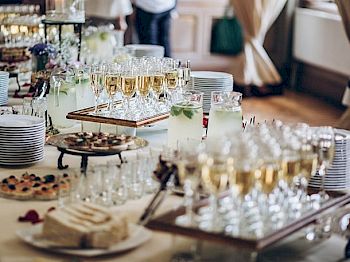 The image shows a buffet table set with glasses of champagne, plates of appetizers, pitchers of lemon water, and a cake, ready for guests.