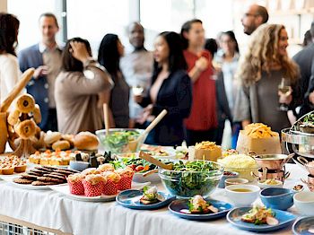 People are mingling at an event with a table of assorted foods including salads, pastries, and small dishes, in the foreground.