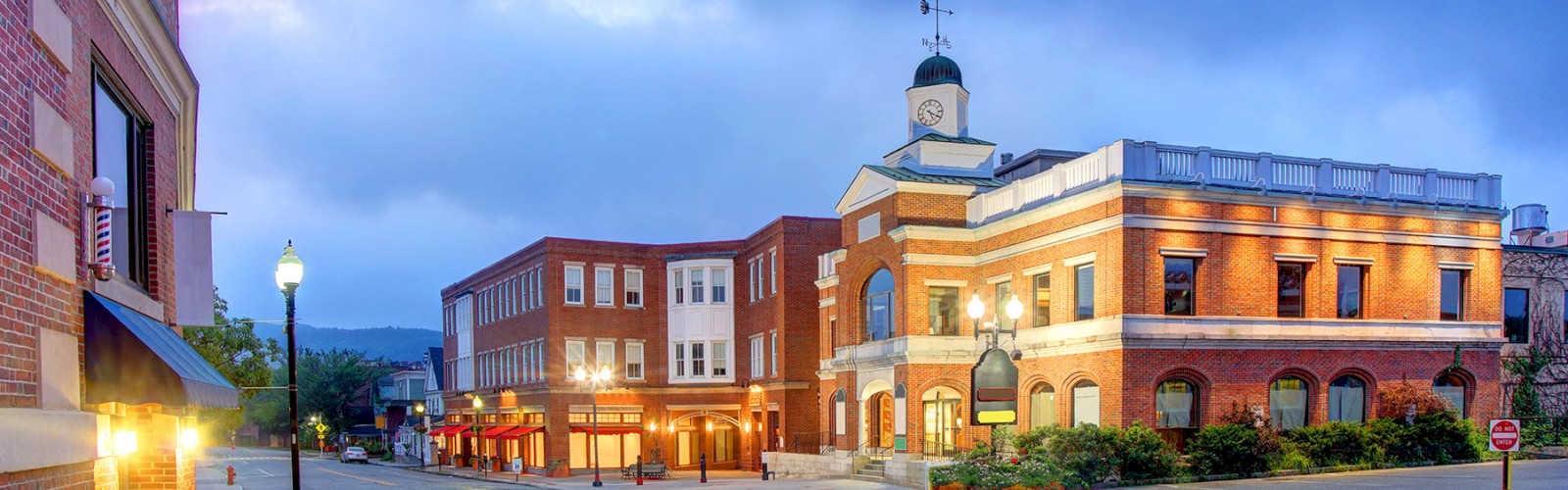 A picturesque small town street with brick buildings, including a clock tower, under a dusky sky with street lighting and greenery.