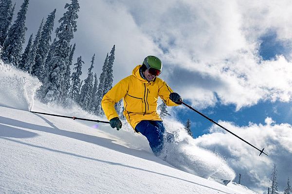A skier in a yellow jacket and green helmet is carving through powdery snow on a sunny day, with snow-covered trees in the background.