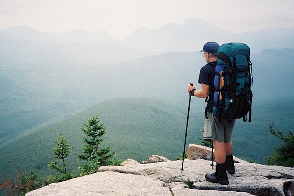 A hiker stands on a rocky ledge with trekking poles, wearing a large backpack, overlooking a foggy, mountainous landscape.