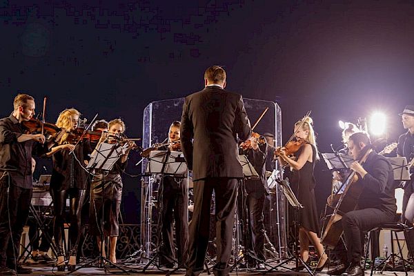 The image shows an orchestra performing on stage, with musicians playing string instruments and a conductor leading them, under bright stage lights.