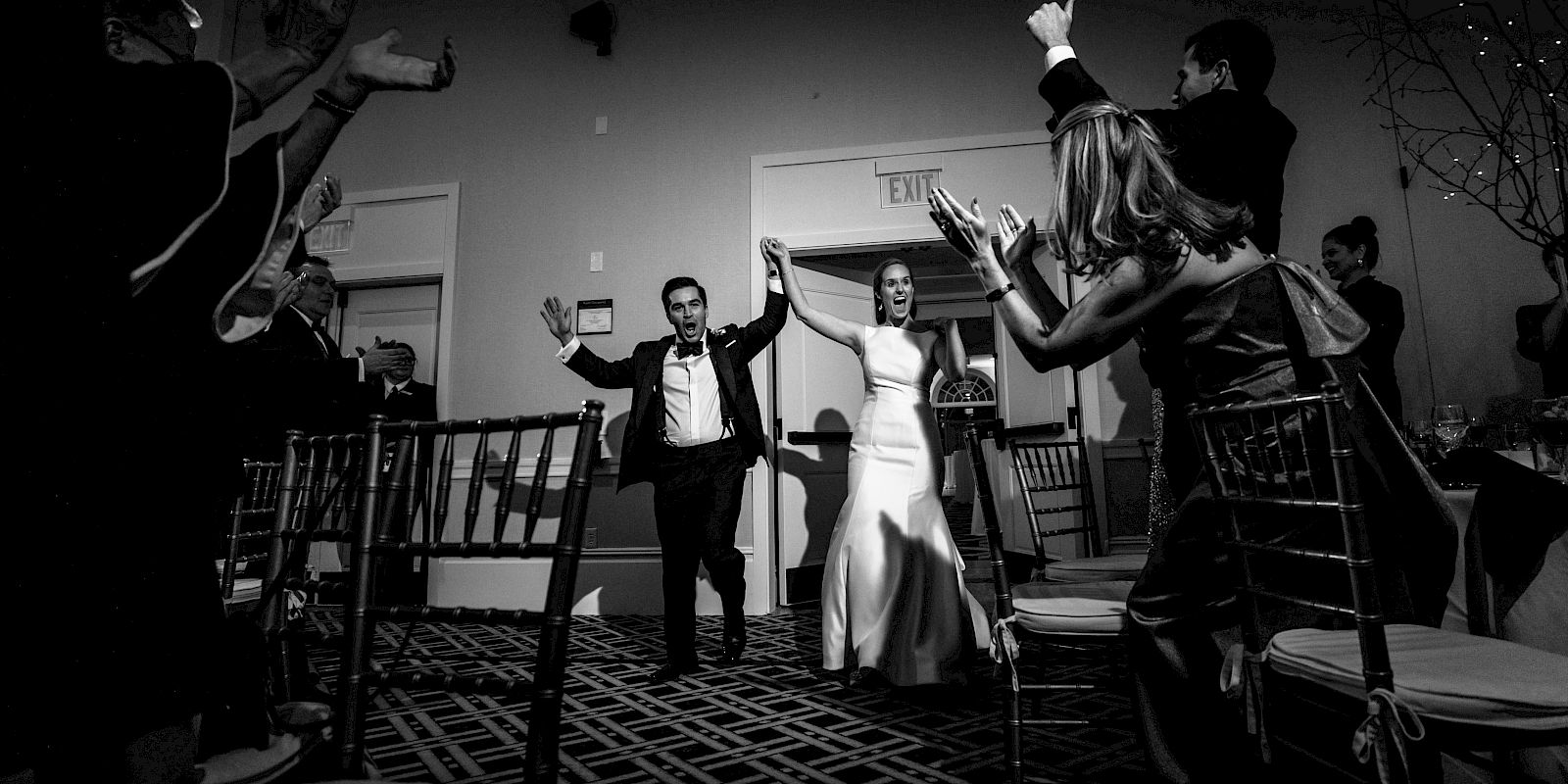 A newlywed couple is joyfully entering a reception hall, surrounded by cheering guests, all dressed formally with raised arms, in a black and white photo.