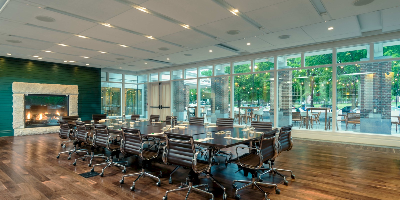 The image shows a modern conference room with a long table, office chairs, a fireplace, large windows, and an outdoor view.