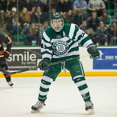 A hockey player in a green and white uniform is on the ice in an arena, holding a stick, with other players and an audience in the background.