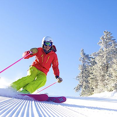 A skier in bright gear is making turns on a snowy slope surrounded by snow-covered trees, under a clear blue sky.