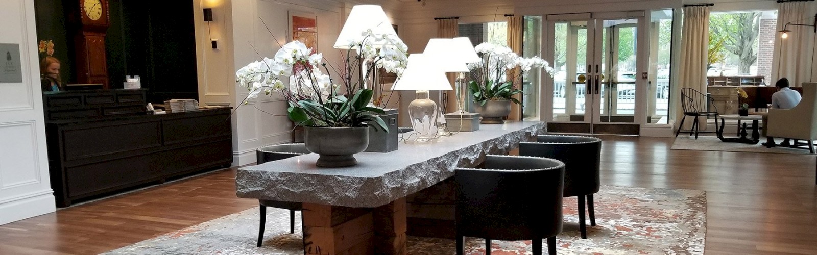A modern lobby with a long stone table, potted orchids, table lamps, chairs, and a cozy seating area in the background.