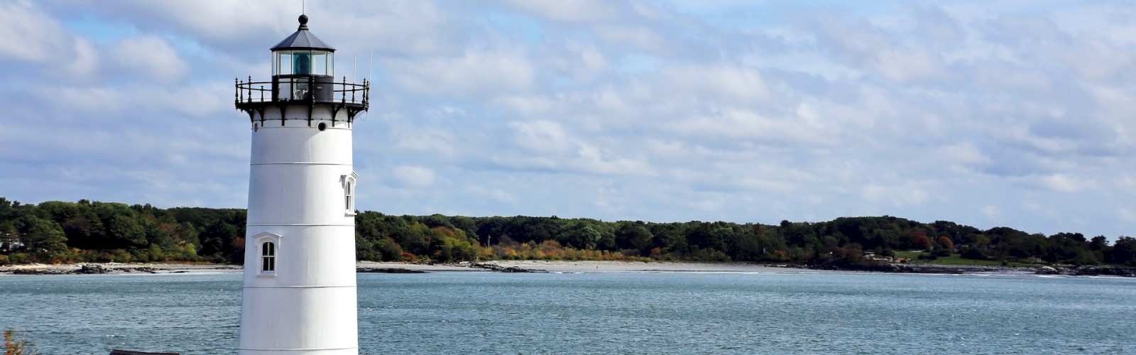 The image shows a white lighthouse situated on rocky terrain near a body of water, with a blue sky and some clouds overhead, and trees in the background.