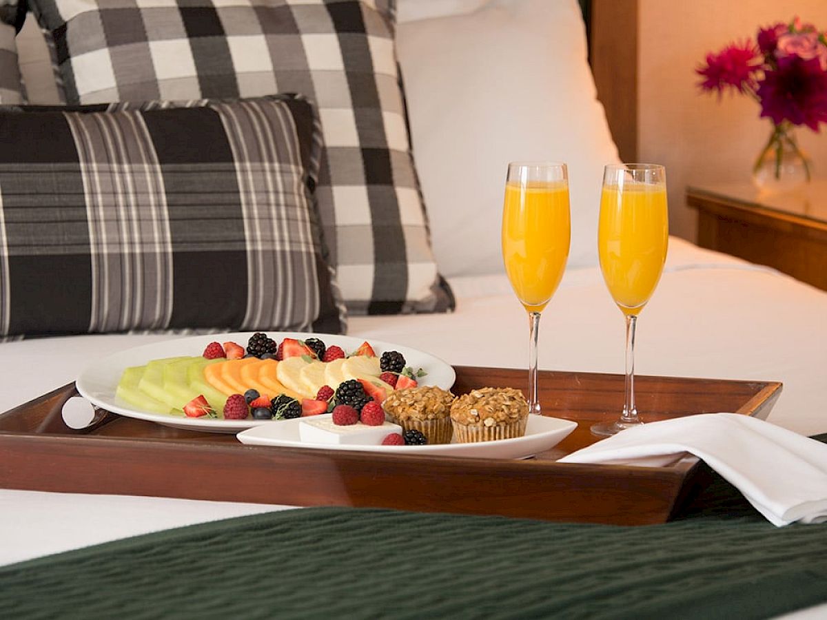 A breakfast tray with fresh fruit, muffins, and two glasses of orange juice is placed on a bed with plaid pillows and a green blanket.